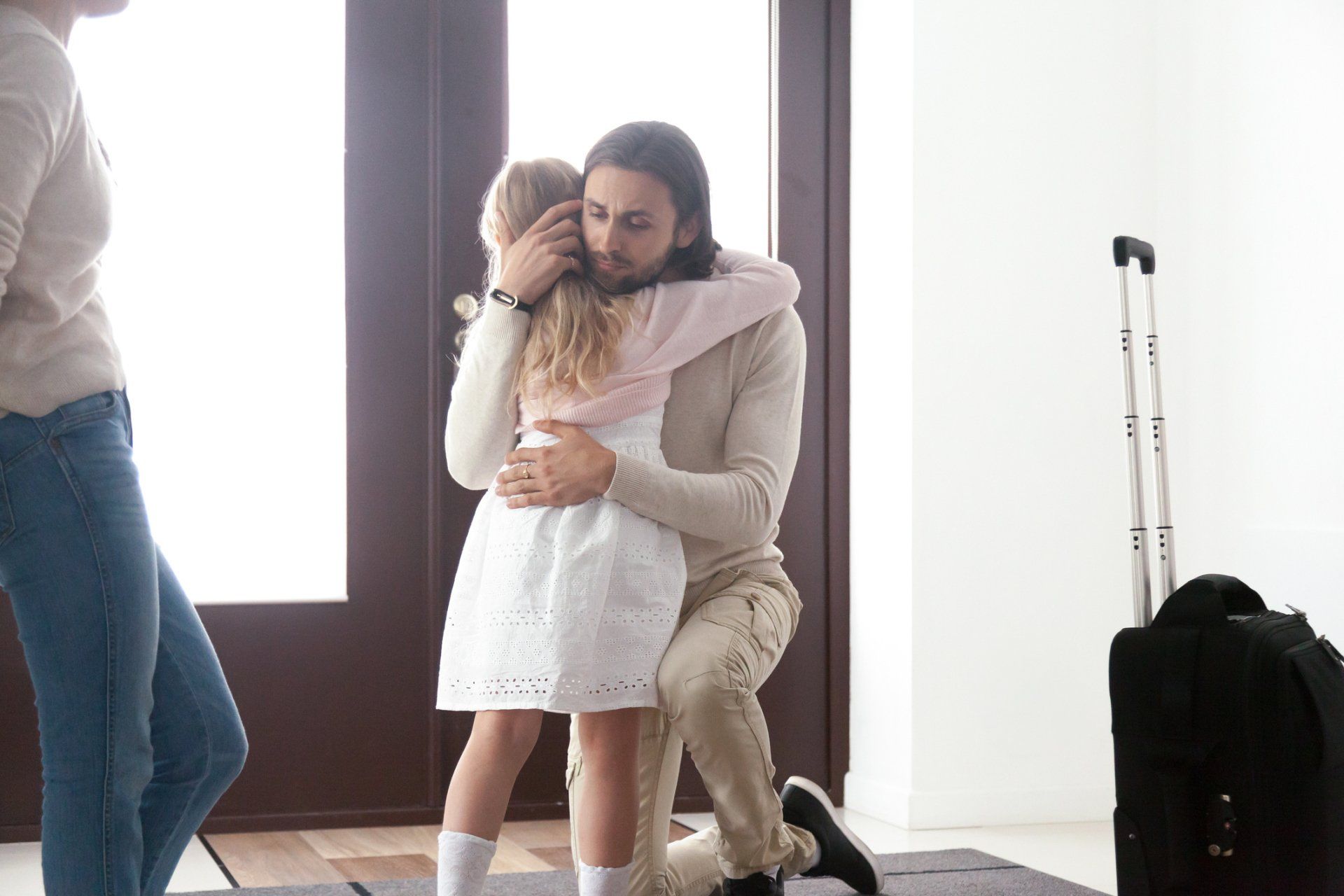 Man kneeling, hugging a child in a doorway, suitcase nearby; woman watches.
