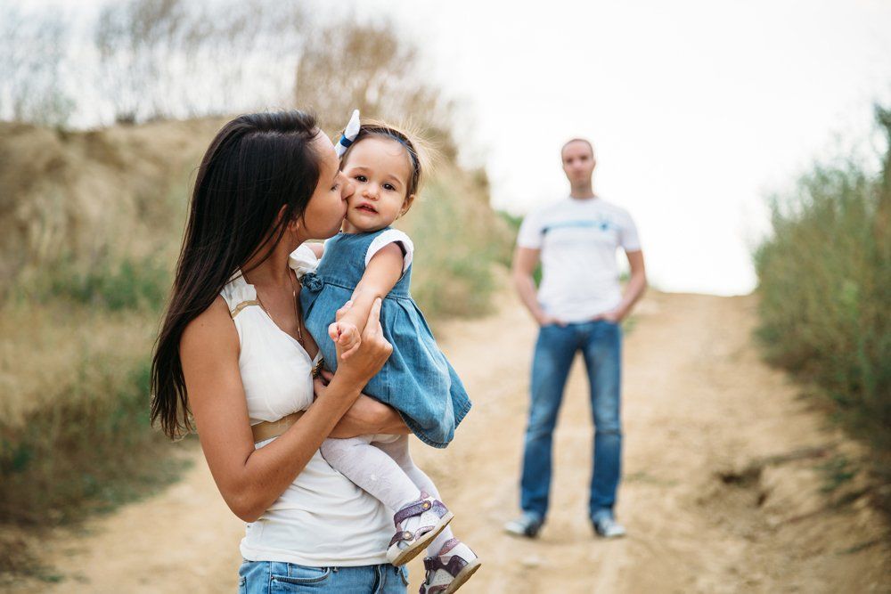 Woman kissing a child; man stands in background on dirt path. Outdoors, neutral tones.