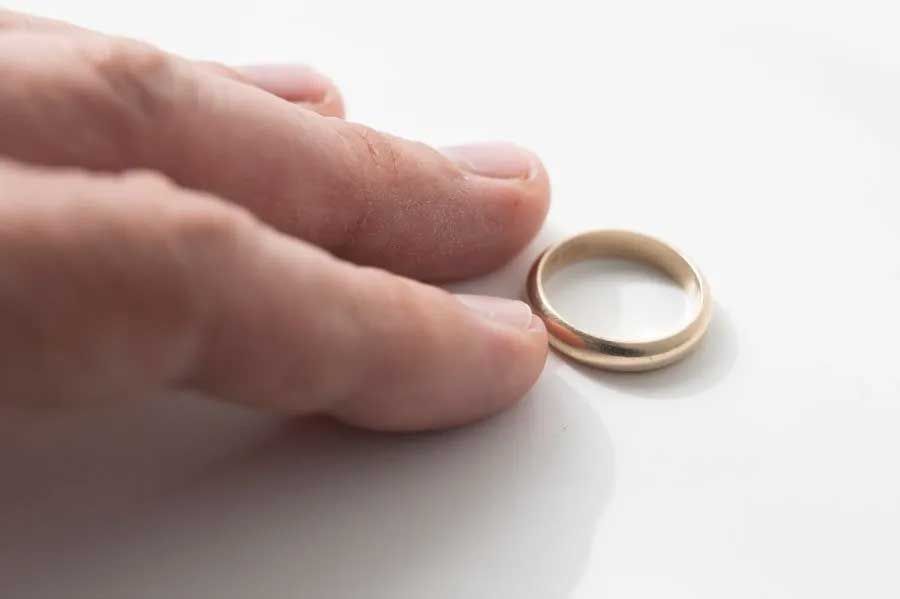 Hand reaching for a gold wedding band on a white surface.