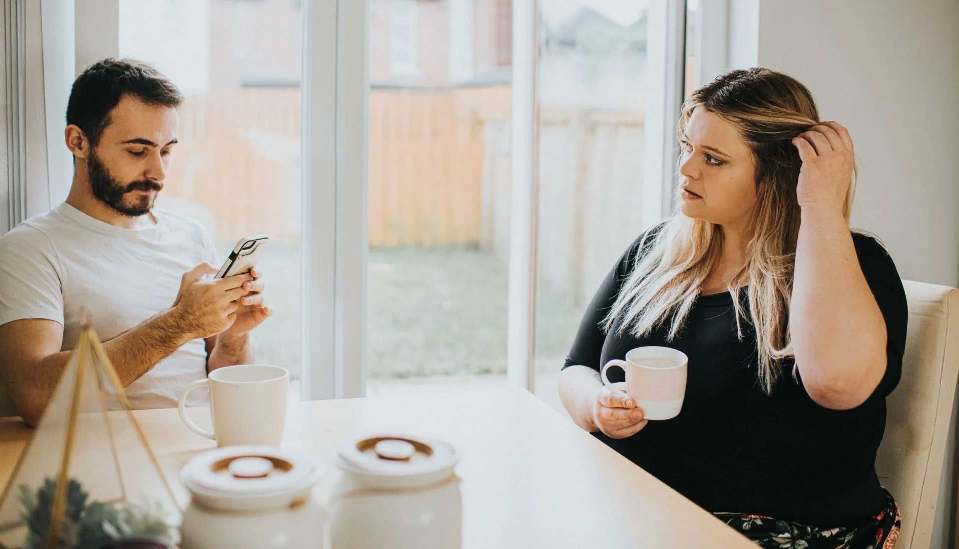 Man on phone, woman holding mug. Both at table in a bright room.
