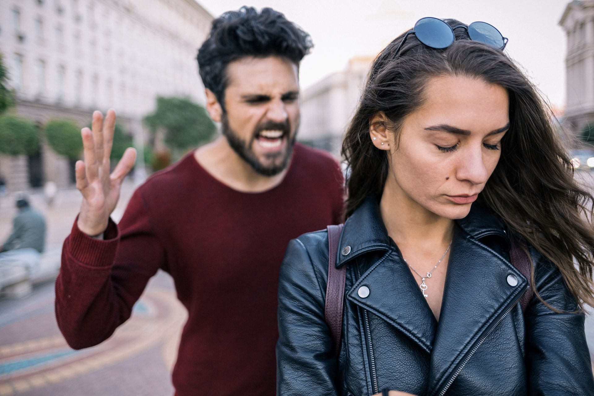 Man yelling behind woman with downward gaze; outdoor setting.