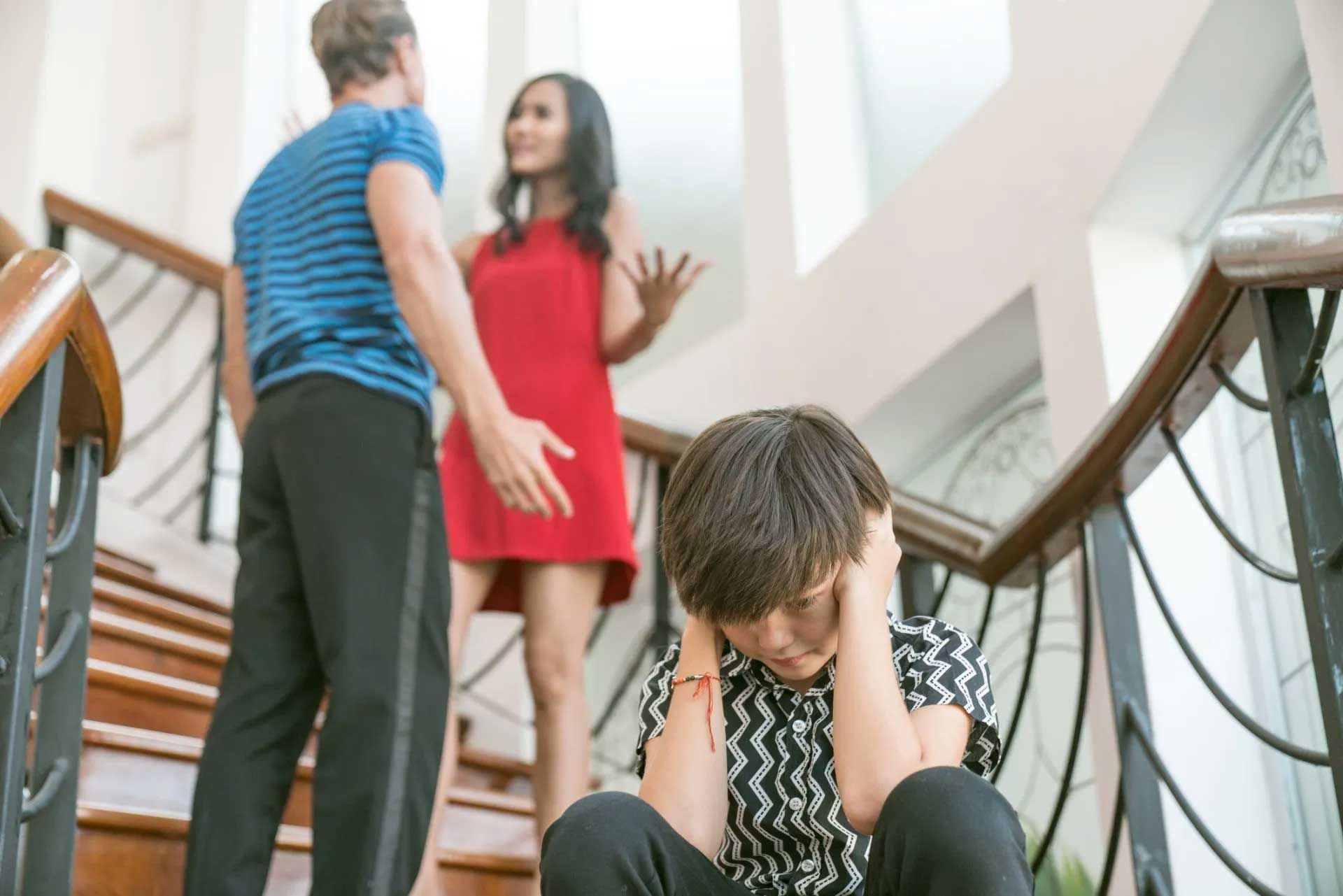 A child covers their ears on stairs as parents argue nearby in a home setting.