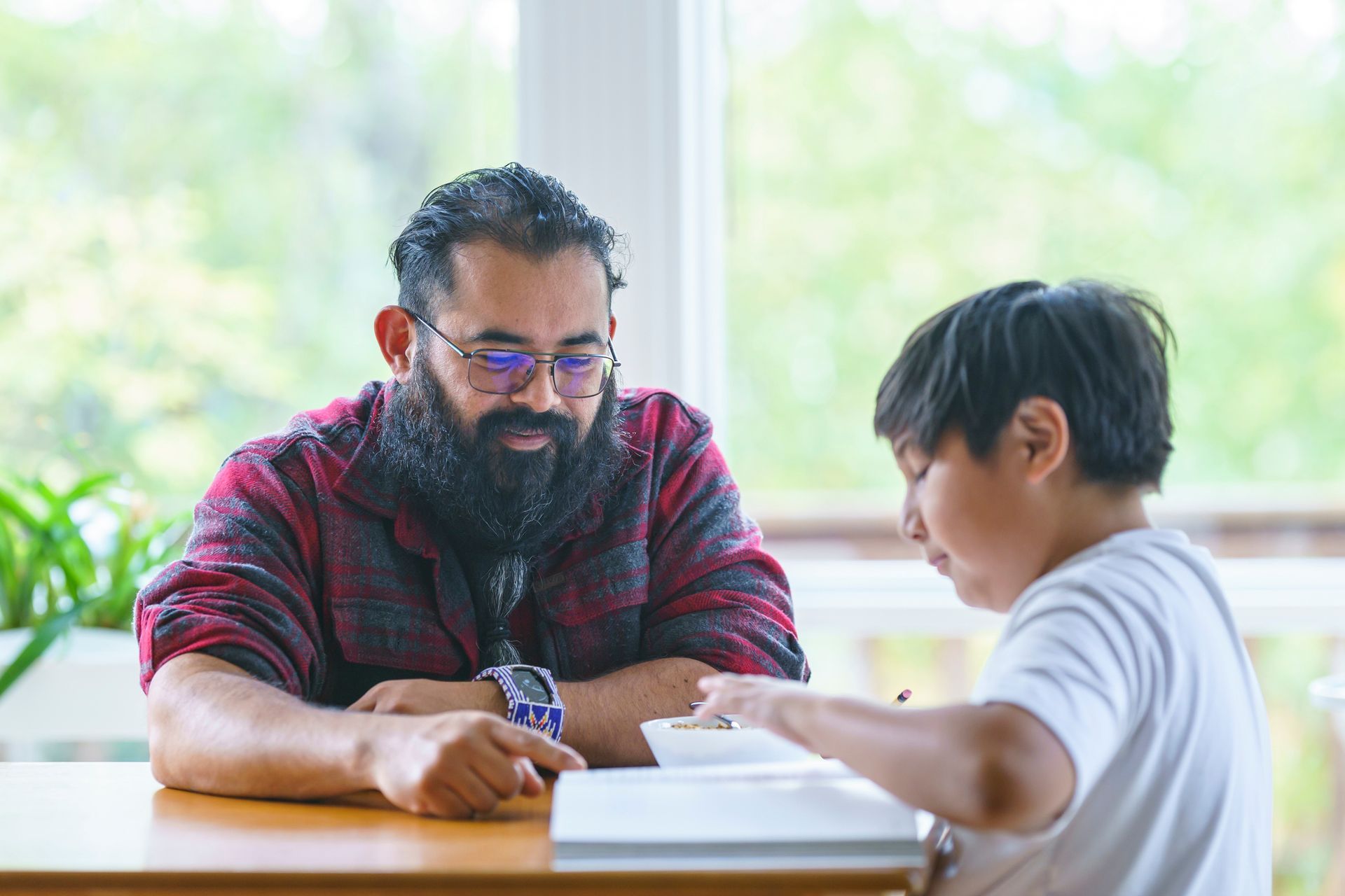 Man with glasses points to book while boy writes, at table near window.