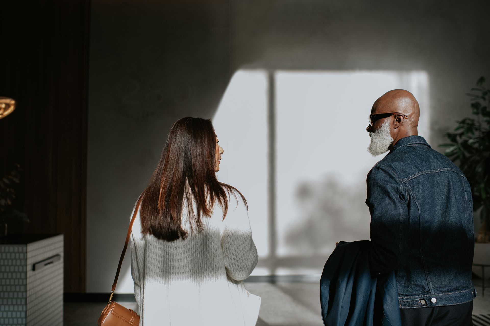 Woman and man walking toward a bright window. The man has a white beard and denim jacket.