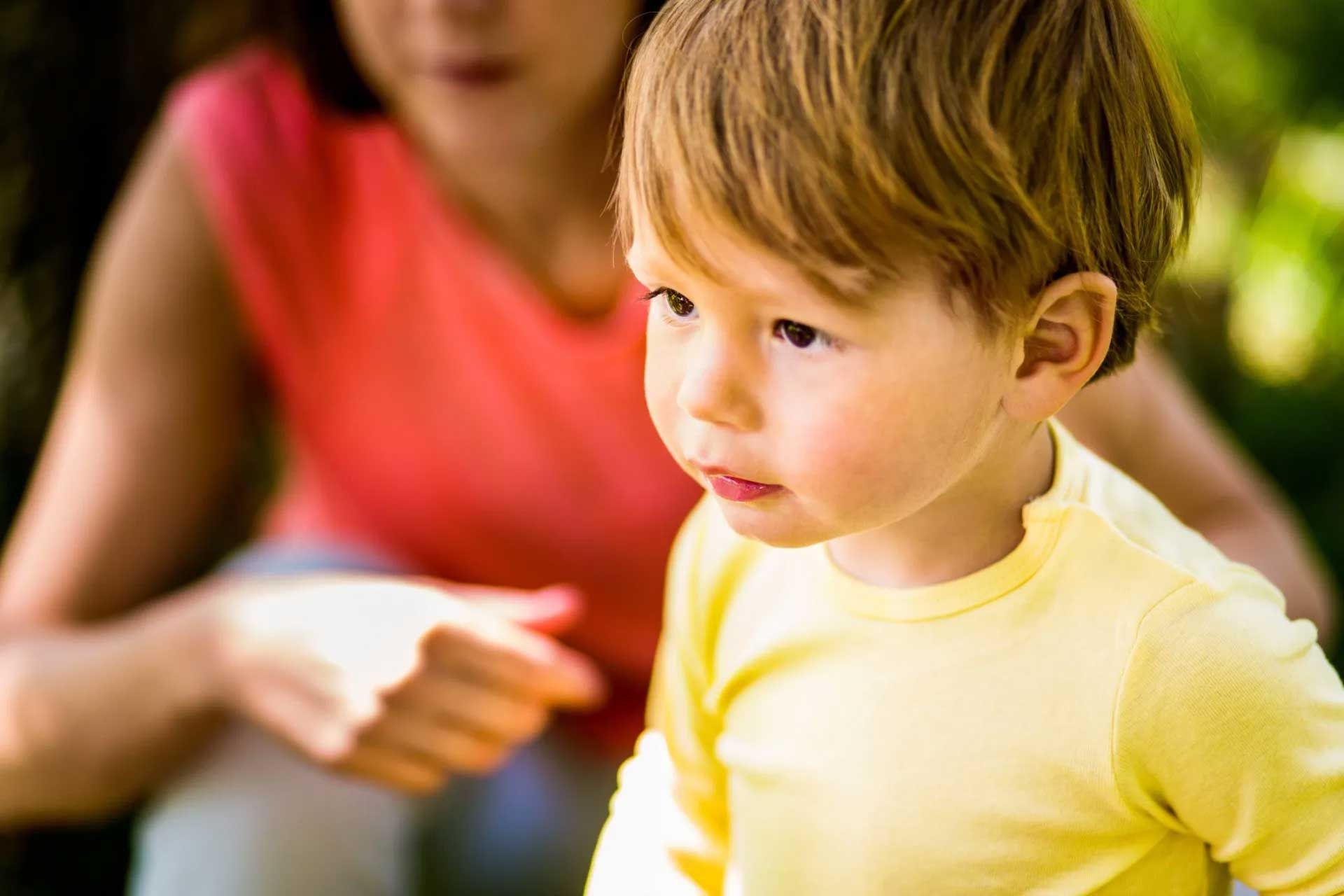 Young child in yellow shirt looking away, with a person in pink out of focus behind him.