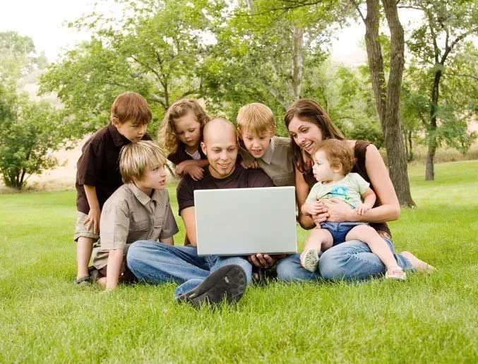 Family of seven gathered outdoors, looking at a laptop computer on a grassy lawn.