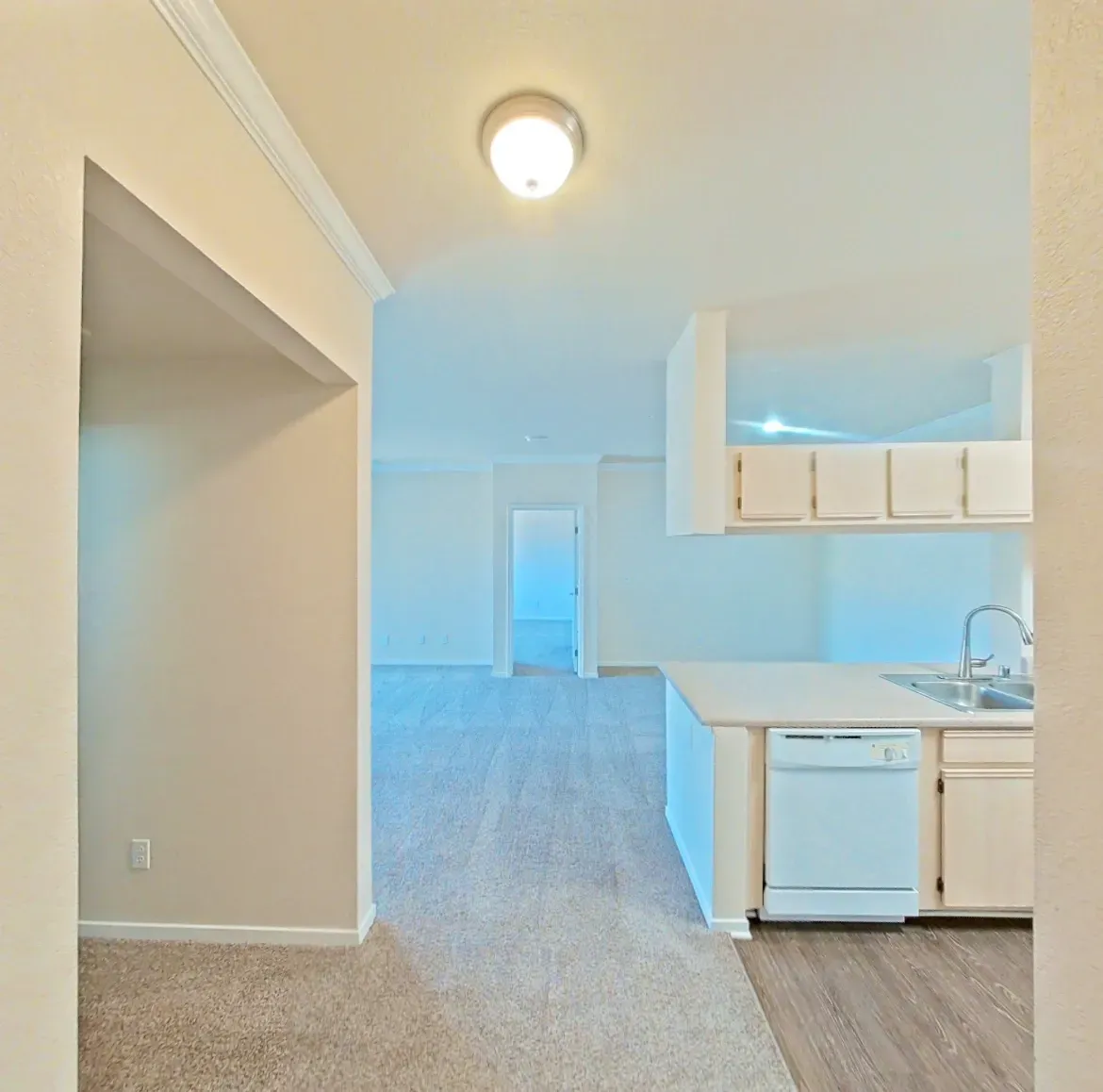 View of a neutral-toned apartment kitchen and living area with carpet and wood laminate flooring.