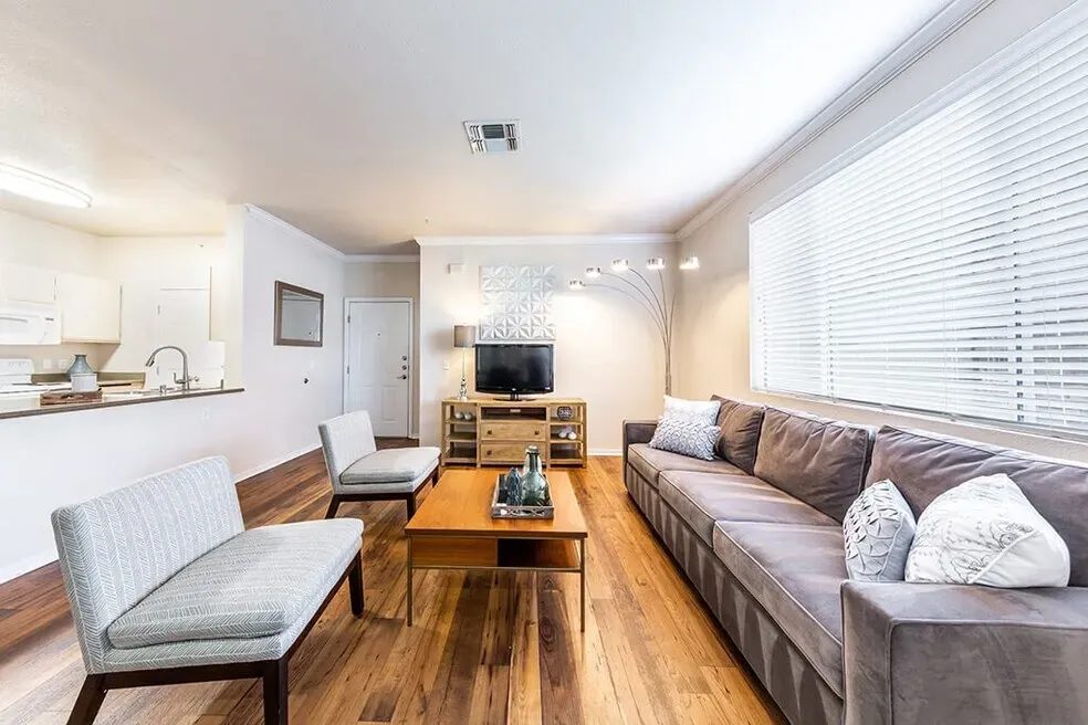 Living room with plush sofa, coffee table, and accent chairs. Kitchenette visible in background.