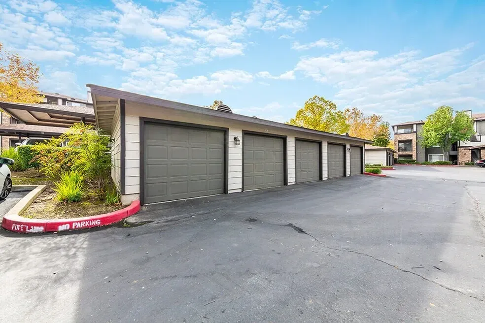 Garages with gray doors in a row, with covered parking and apartment buildings in the background.
