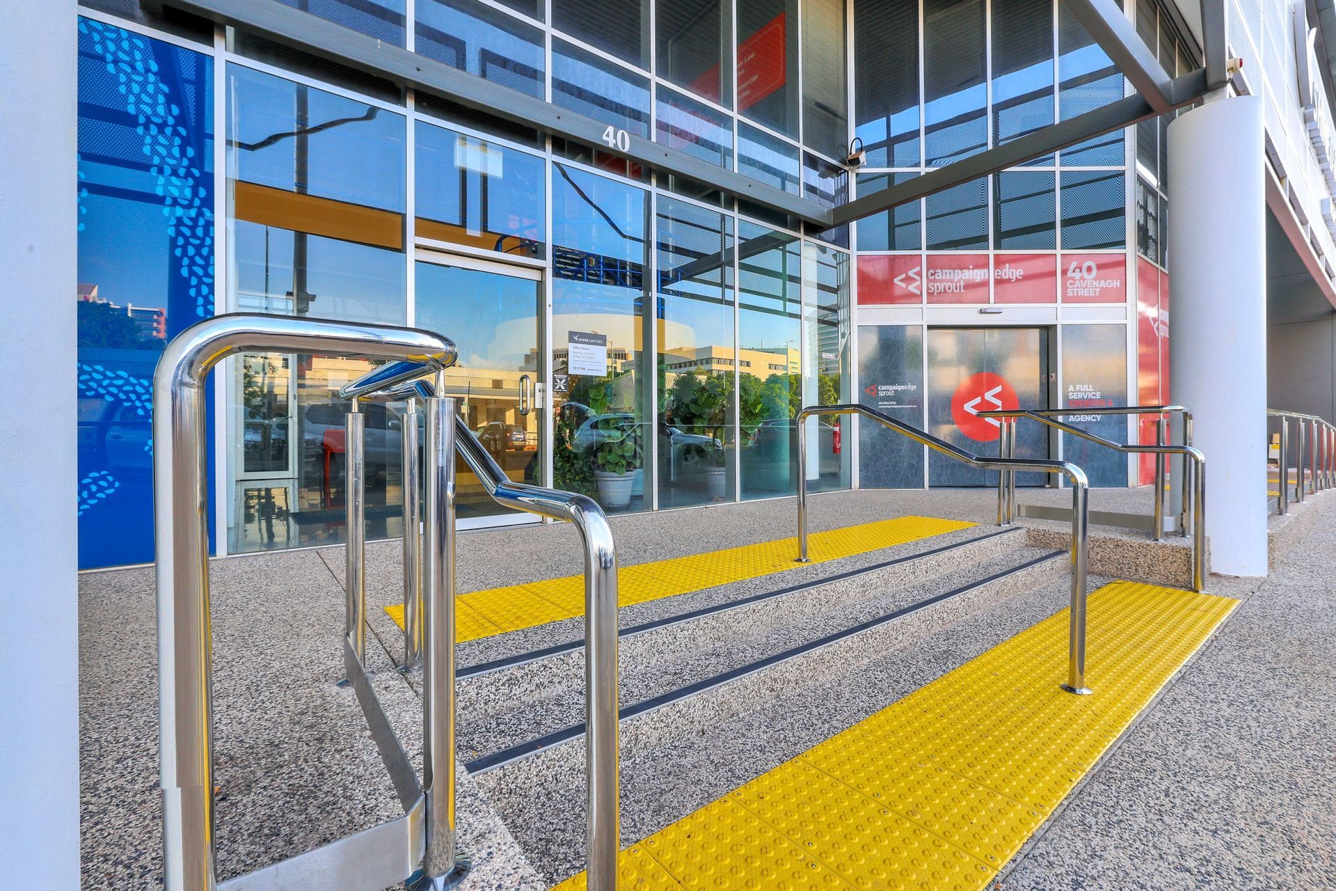 Building entrance with concrete stairs, metal handrails, and bright yellow tactile paving strips for accessibility. — Invision NT in Coconut Grove, NT