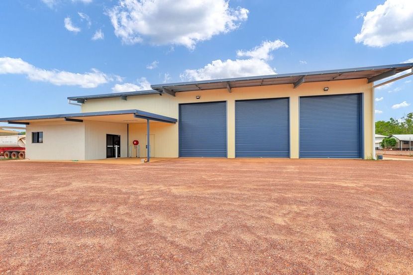 Low beige building with three large blue garage doors on a gravel lot under a partly cloudy sky — Invision NT in Gove, NT