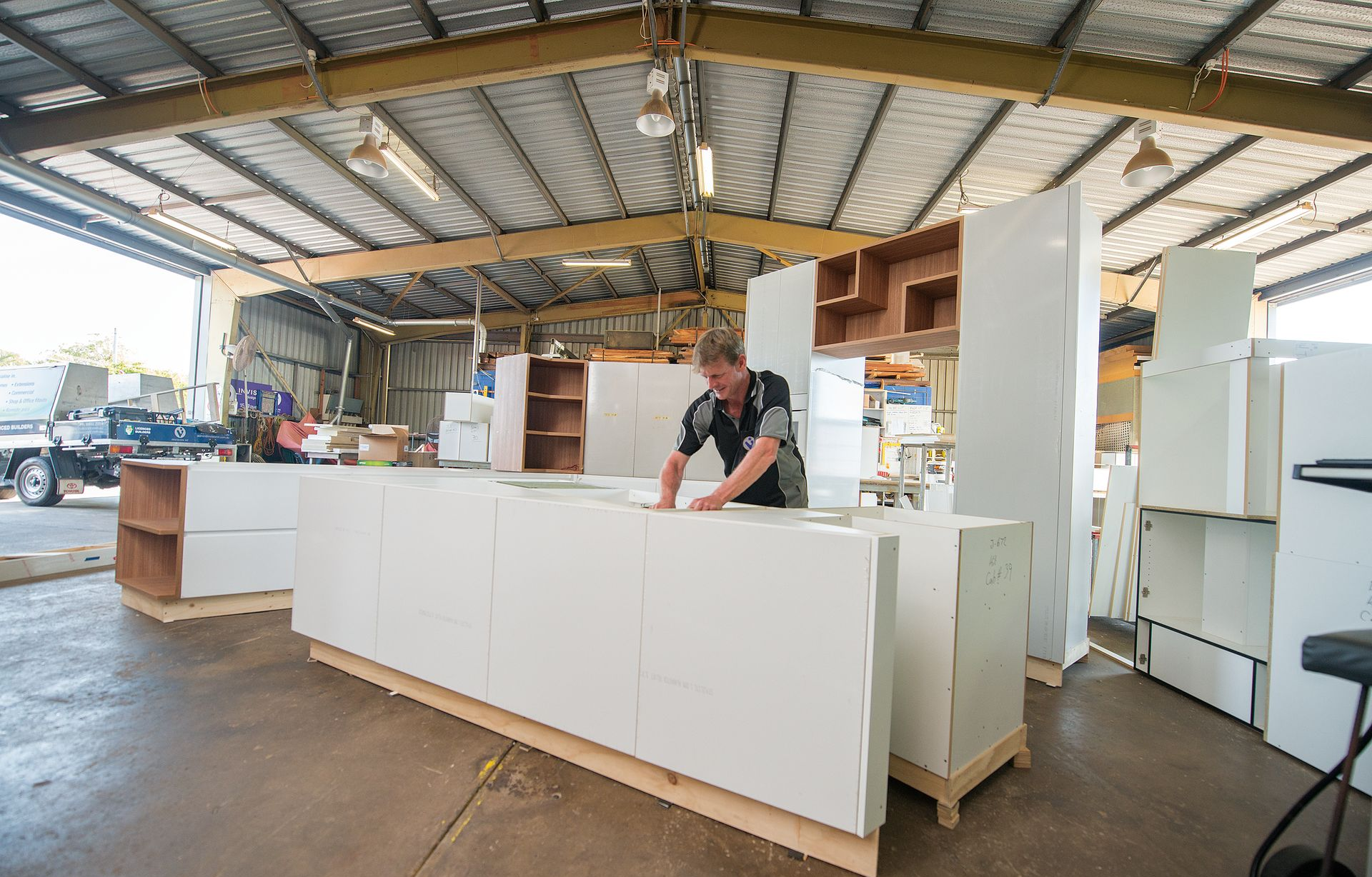 Man assembling white cabinets in a workshop under a metal roof; other cabinets and tools are visible — Invision NT in Coconut Grove, NT