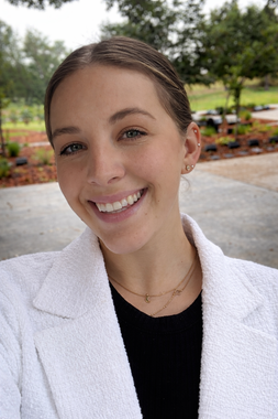 Smiling person in a white blazer standing outdoors in a park setting