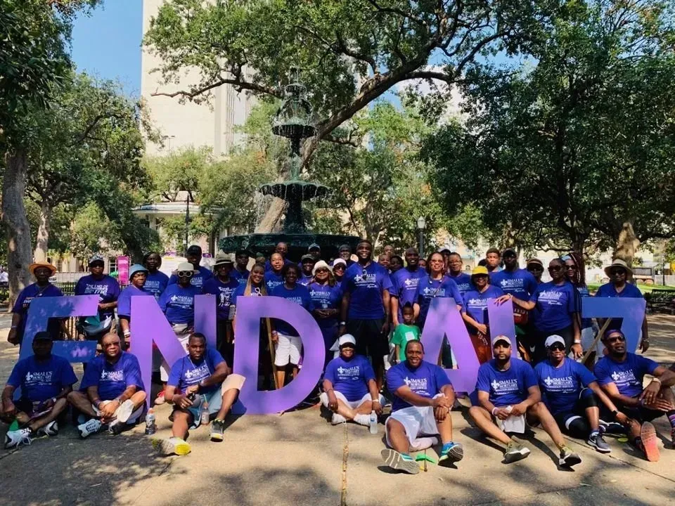 A group poses in front of a fountain, holding large purple letters that spell