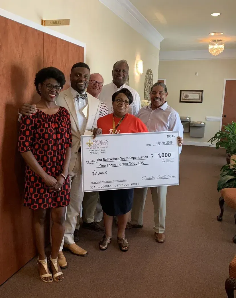 Six people stand in a hallway holding a large commemorative donation check, smiling while posing for a group photo.