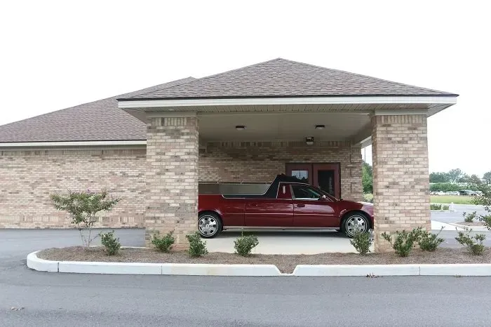 A red hearse parked under the covered entrance of a brick funeral home building.