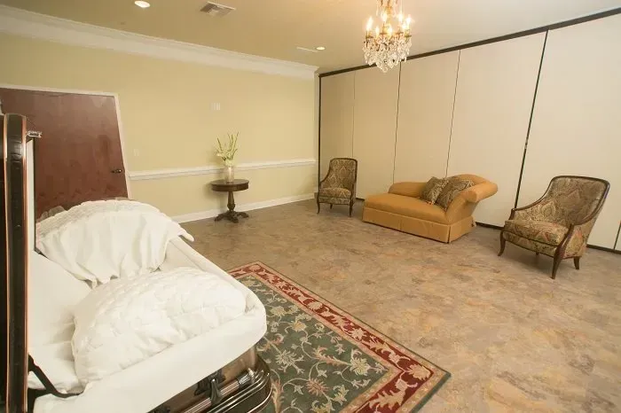 A funeral viewing room with a casket in the foreground, two armchairs, and a lounge chair on a patterned rug.