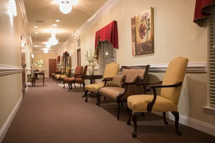 A long hallway with neutral walls, carpeted floors, elegant yellow armchairs, and framed art decor.