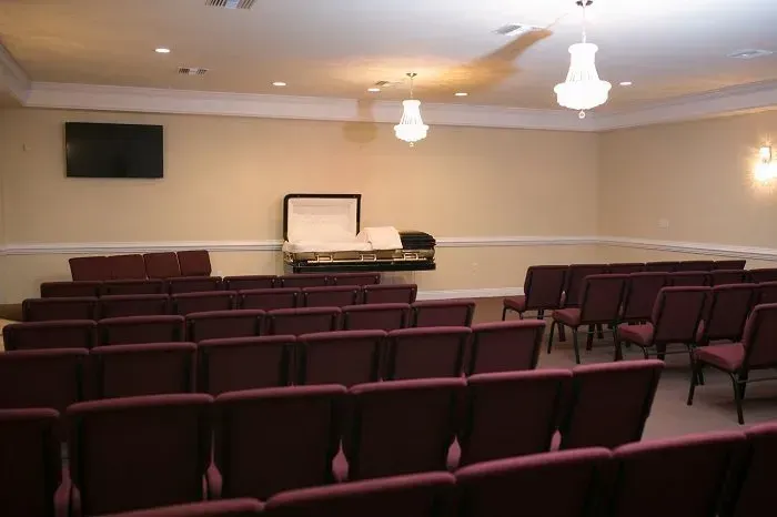 A funeral home viewing room with rows of maroon chairs facing an open casket on a stand beneath a television screen.