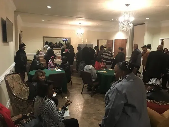 A group of people gathers in a well-lit community hall with green-clothed tables, chairs, and two large crystal chandeliers.