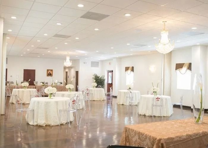 A bright, spacious event hall with round tables covered in white linens, elegant chandeliers, and a tan rectangular table.