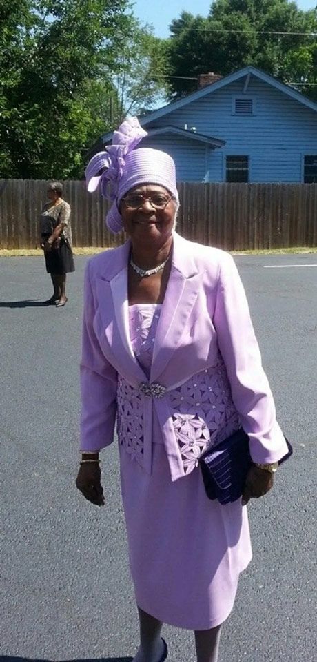 Irene J. Small wearing a light purple suit and matching hat with a large bow & holding a dark clutch.