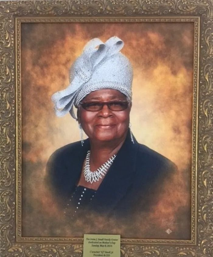 Framed portrait of Irene J. Small wearing a light blue hat, glasses, and a necklace against a tan background.