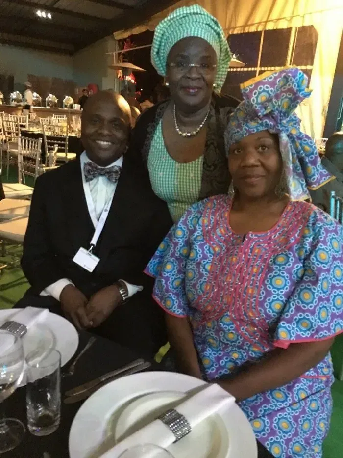 Three people smile at a table with plates and glasses in a decorated indoor venue.