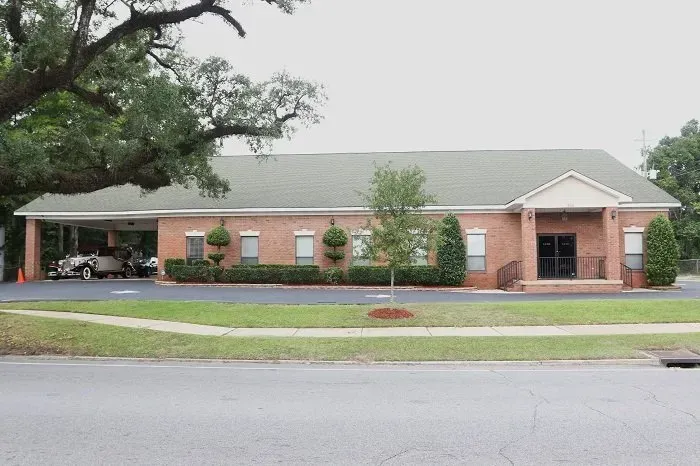 A brick building with a green roof, arched driveway, and central entrance, surrounded by trees and a paved lot.