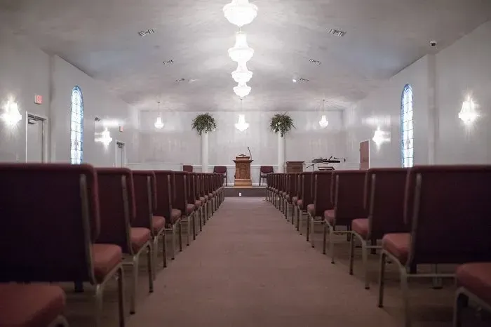 A view down the center aisle of a quiet church sanctuary, featuring rows of red padded chairs and elegant lighting.