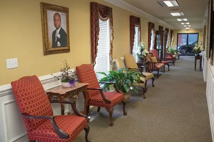 A long, narrow hallway with red upholstered chairs, potted plants, and a portrait hanging on a tan wall.