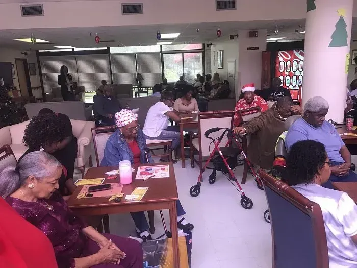 A group of people gather in a community room with tables, chairs, and walkers to participate in a shared activity.