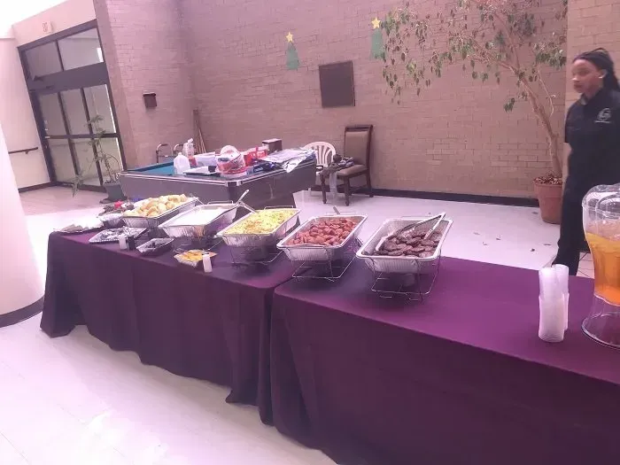 A long table with a purple tablecloth holds several food warming trays in a large room, with a person standing nearby.