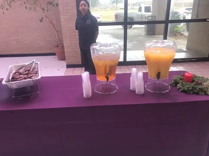 A purple-covered buffet table with a food tray, two beverage dispensers, and a festive wreath near a large window.