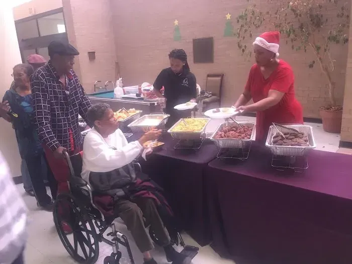 People in a casual, indoor setting serve food buffet-style to an individual seated in a wheelchair.