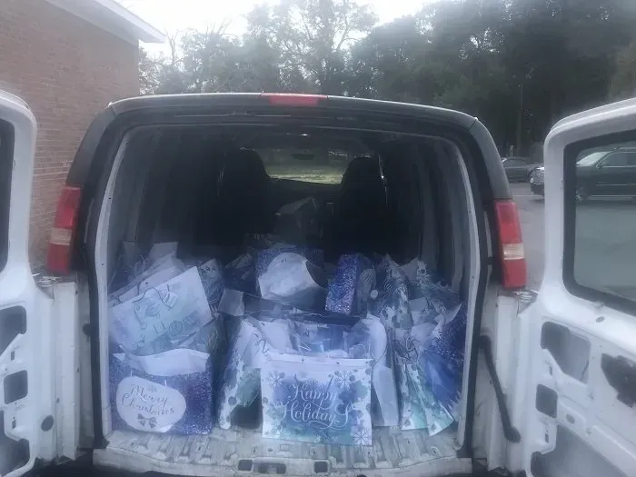 The open back doors of a white van reveal several blue and white gift bags filled with items, parked outdoors.