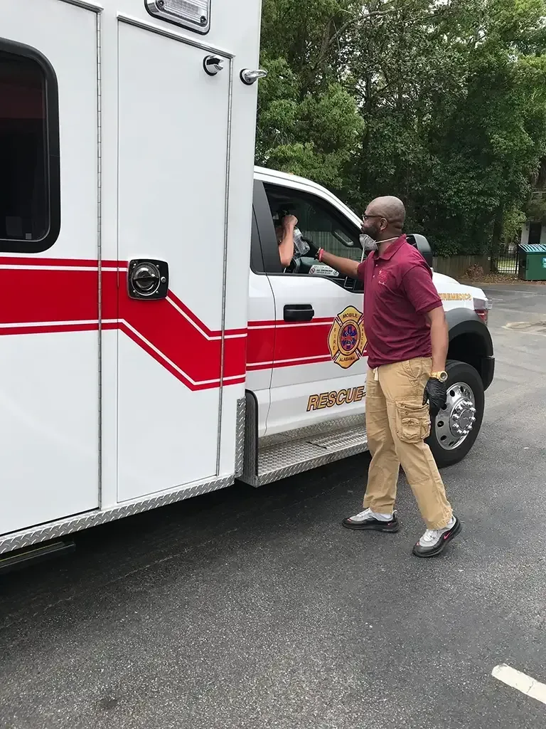 A person in a maroon shirt and tan pants interacts with a driver through the window of a white and red ambulance.