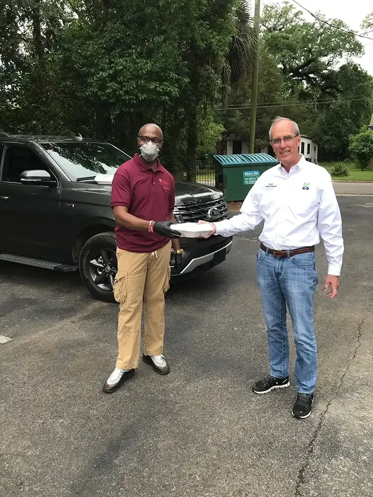 Two people standing outdoors, exchanging a white container in front of a dark vehicle parked near a dumpster.