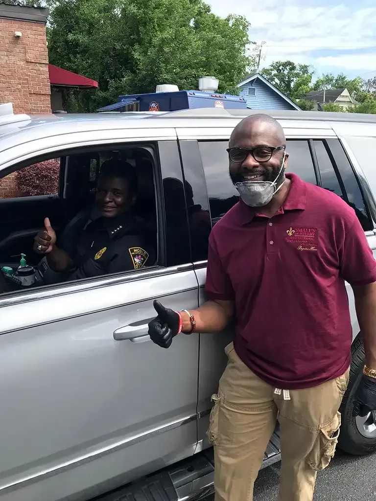 A person in a maroon shirt and mask gives a thumbs-up next to a driver in a law enforcement uniform in a silver SUV.