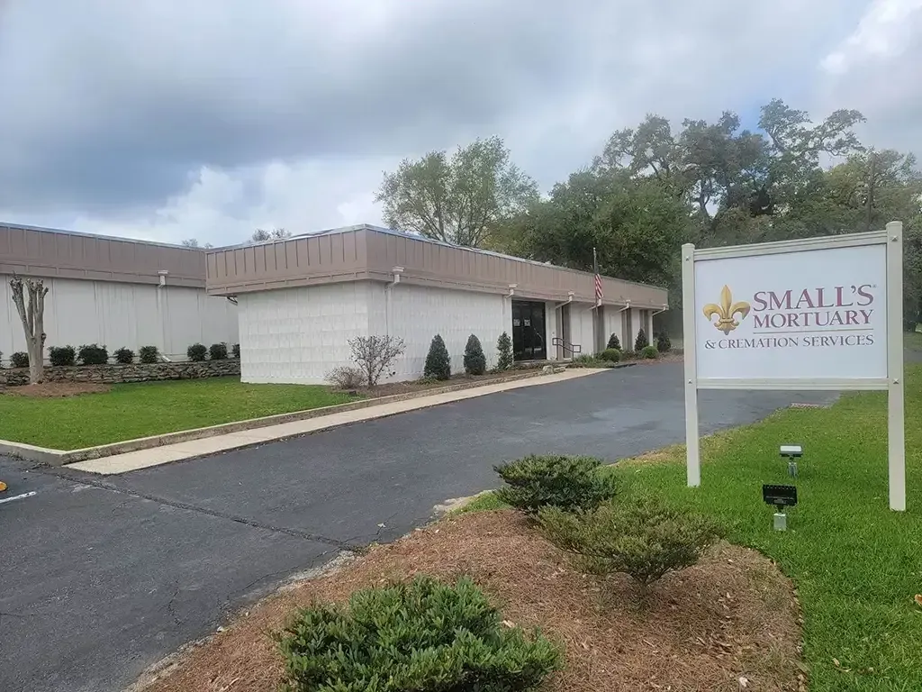 A white building with a brown roof and a sign for Small's Mortuary stands on a grassy lot under a cloudy sky.