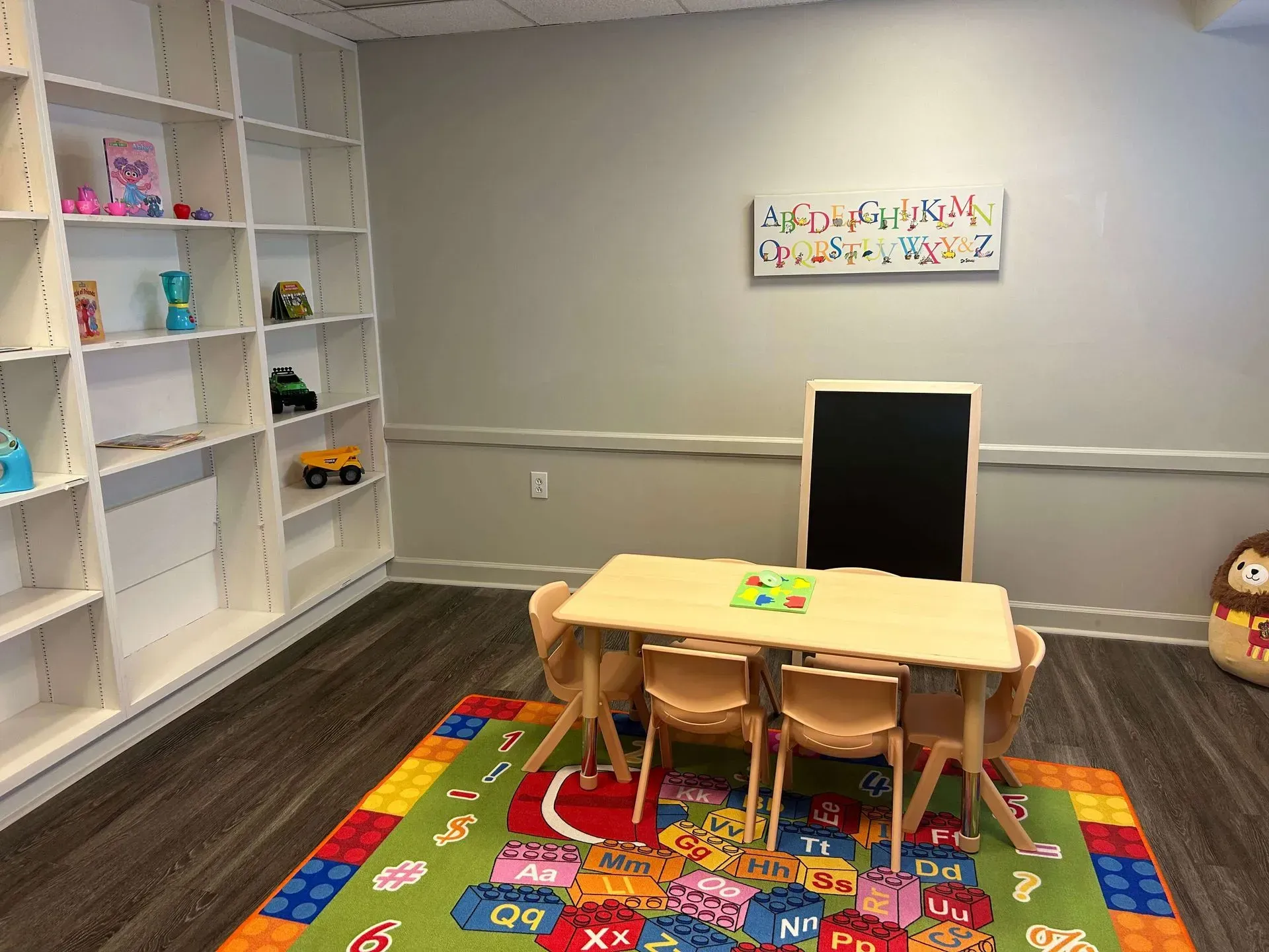 A child's playroom with white shelves, a small wooden table, four chairs, a chalkboard, and an alphabet-patterned rug.