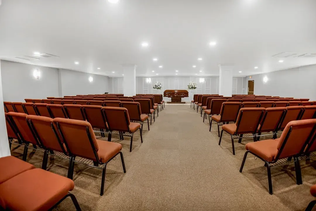 An interior view of a room set up for a service, featuring rows of red padded chairs facing a wooden altar.
