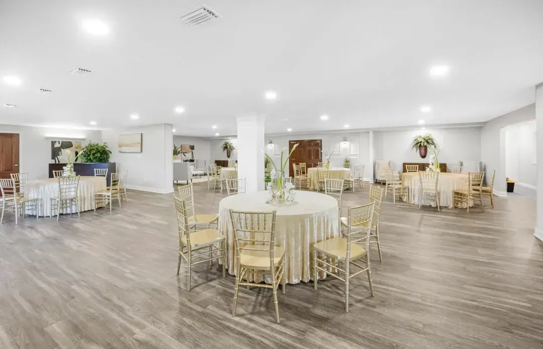 A spacious event hall with beige tablecloth-covered round tables, gold chairs, and light wood-look flooring.