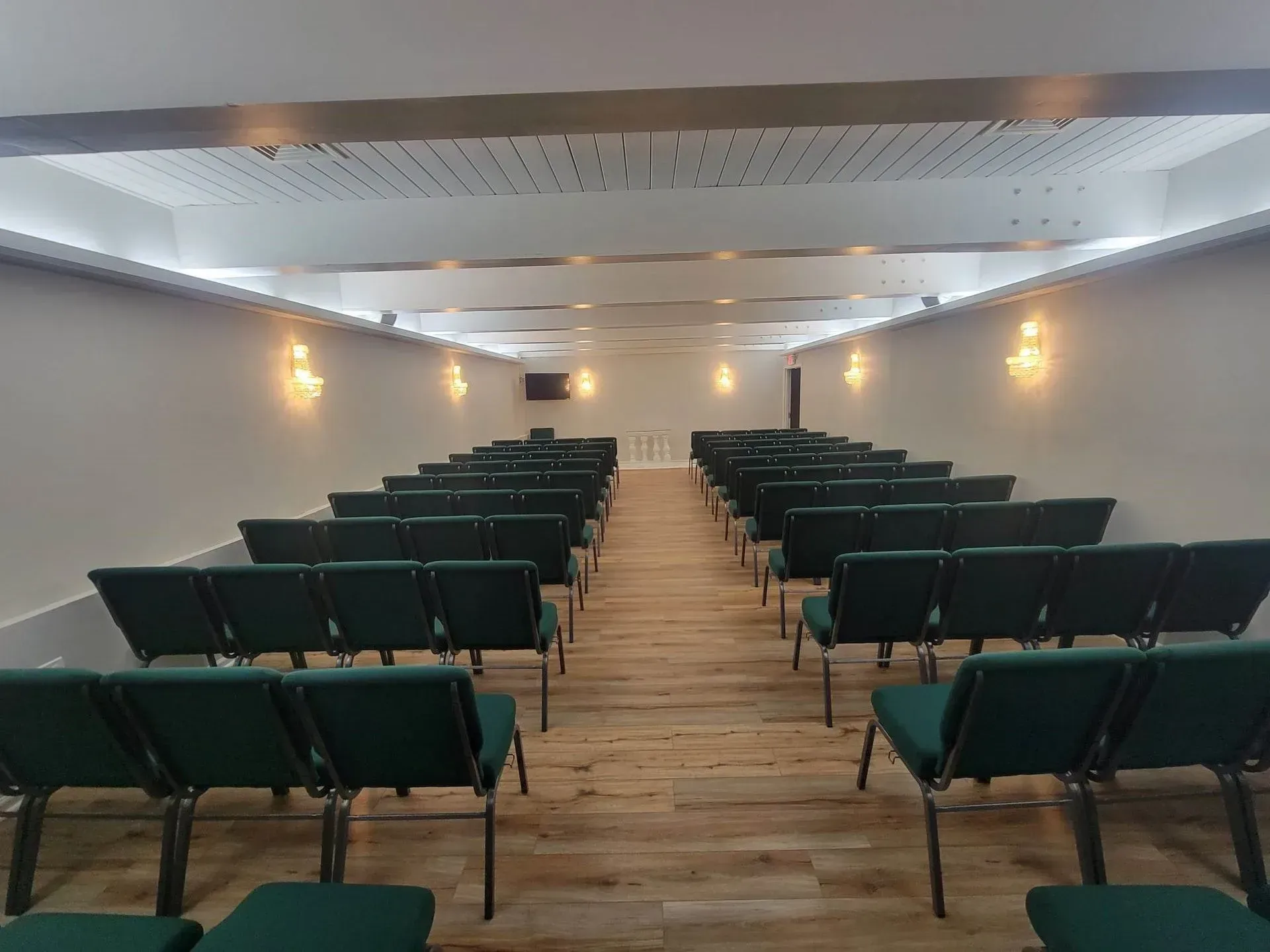 Rows of dark green chairs arranged in a long, rectangular hall with light wood flooring and overhead lights.