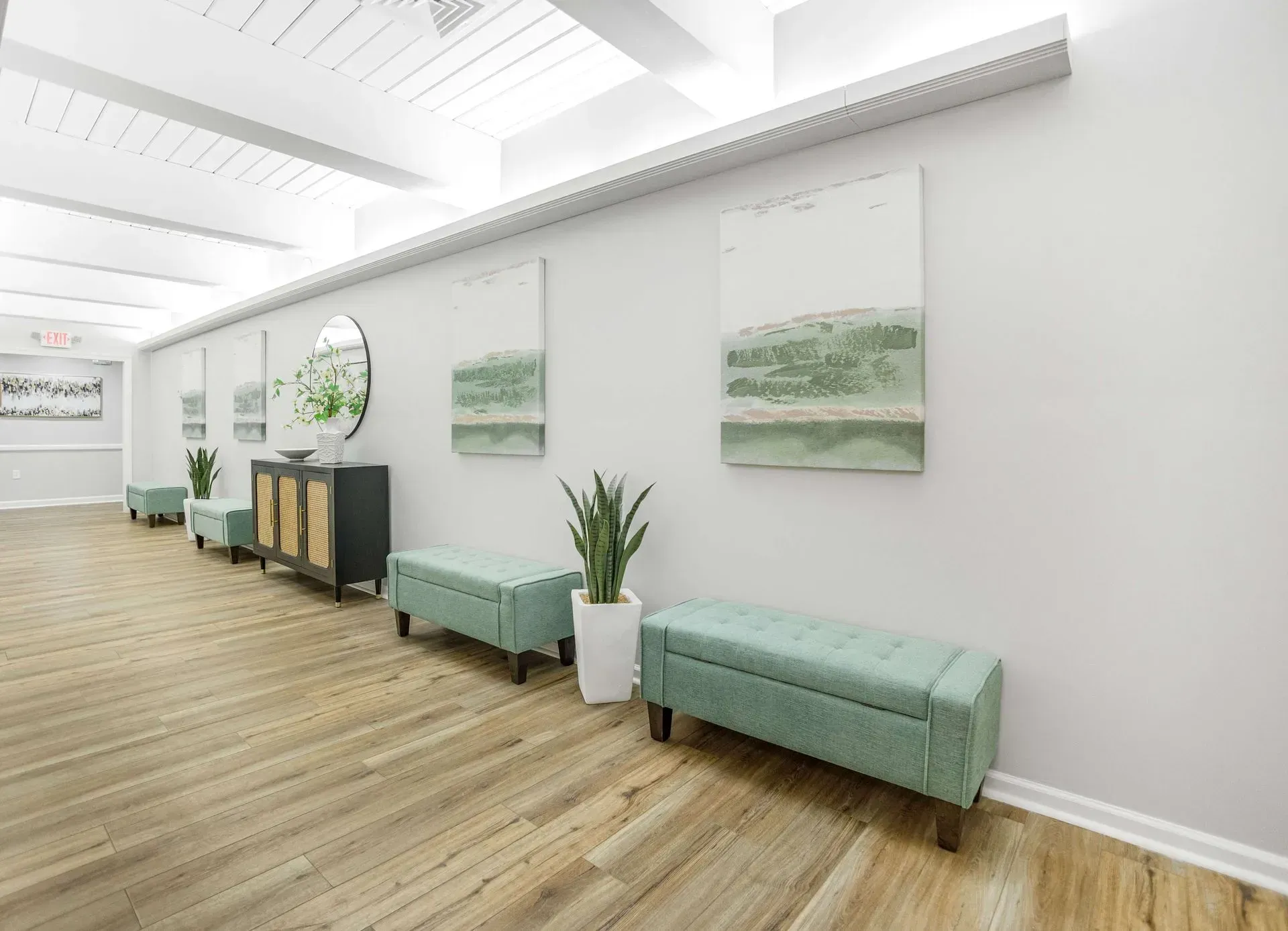 A bright hallway with a light wood floor, two tufted mint-green benches, potted plants, and abstract wall art.