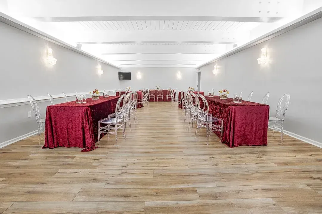 An event hall with two long rows of tables draped in burgundy velvet, clear chairs, and wood-look floors.