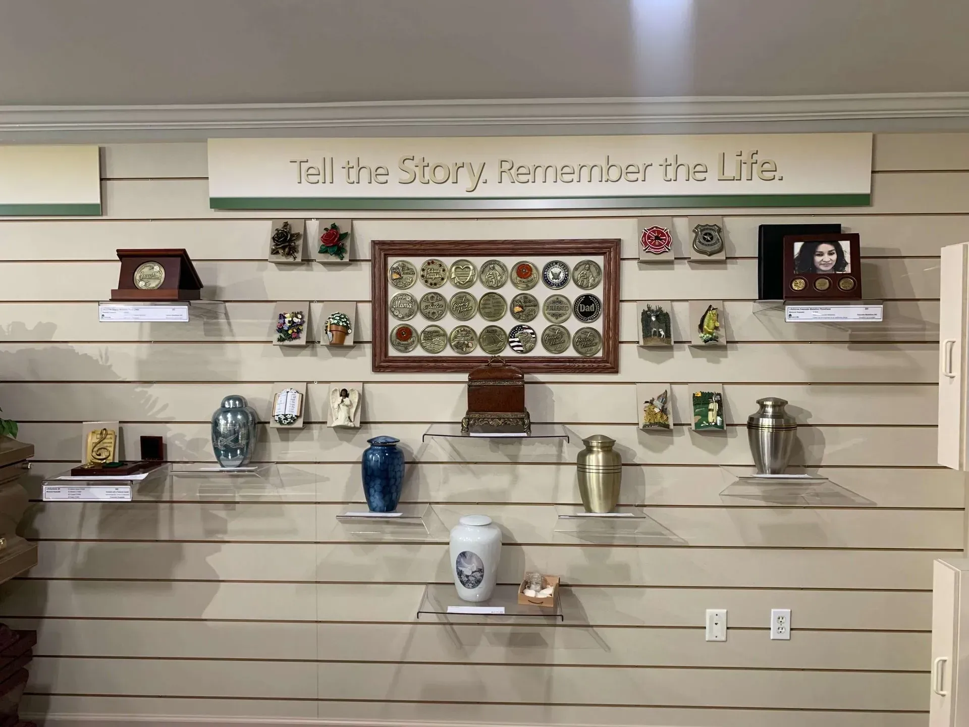 A funeral home wall display features various cremation urns, memorial keepsakes, and a framed collection of mementos.