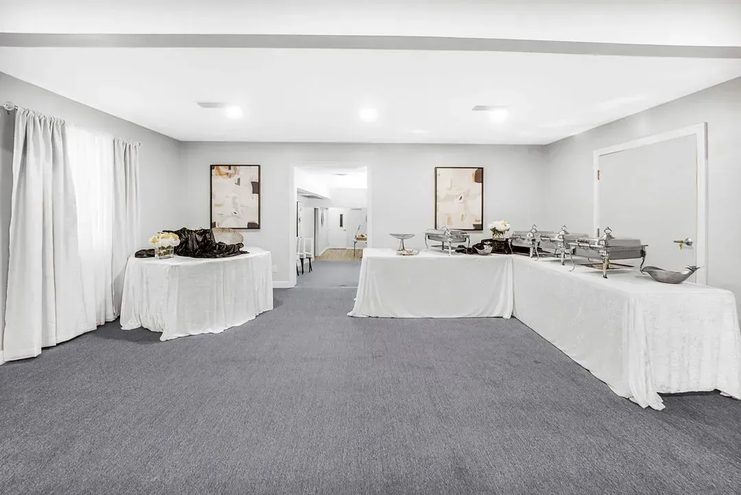 A white-walled room with grey carpet features tables draped in white cloths prepared for a catered event or buffet.