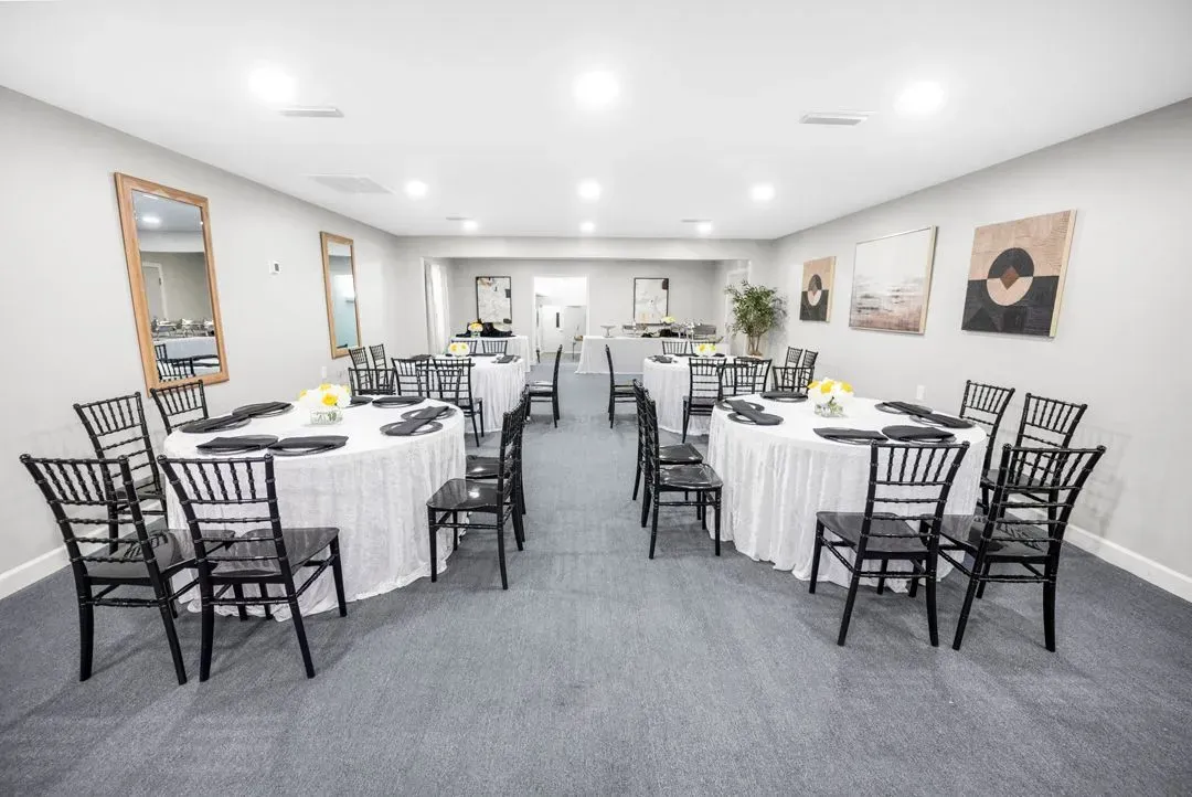 A bright event room with two round tables covered in white linens, surrounded by black chairs on a gray carpet.