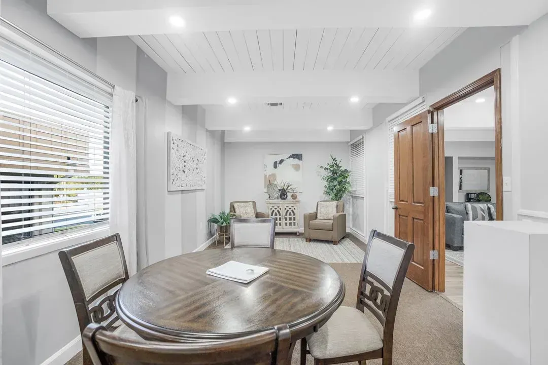 A round wooden dining table and chairs sit in a light-filled room, with armchairs and an open doorway in the background.